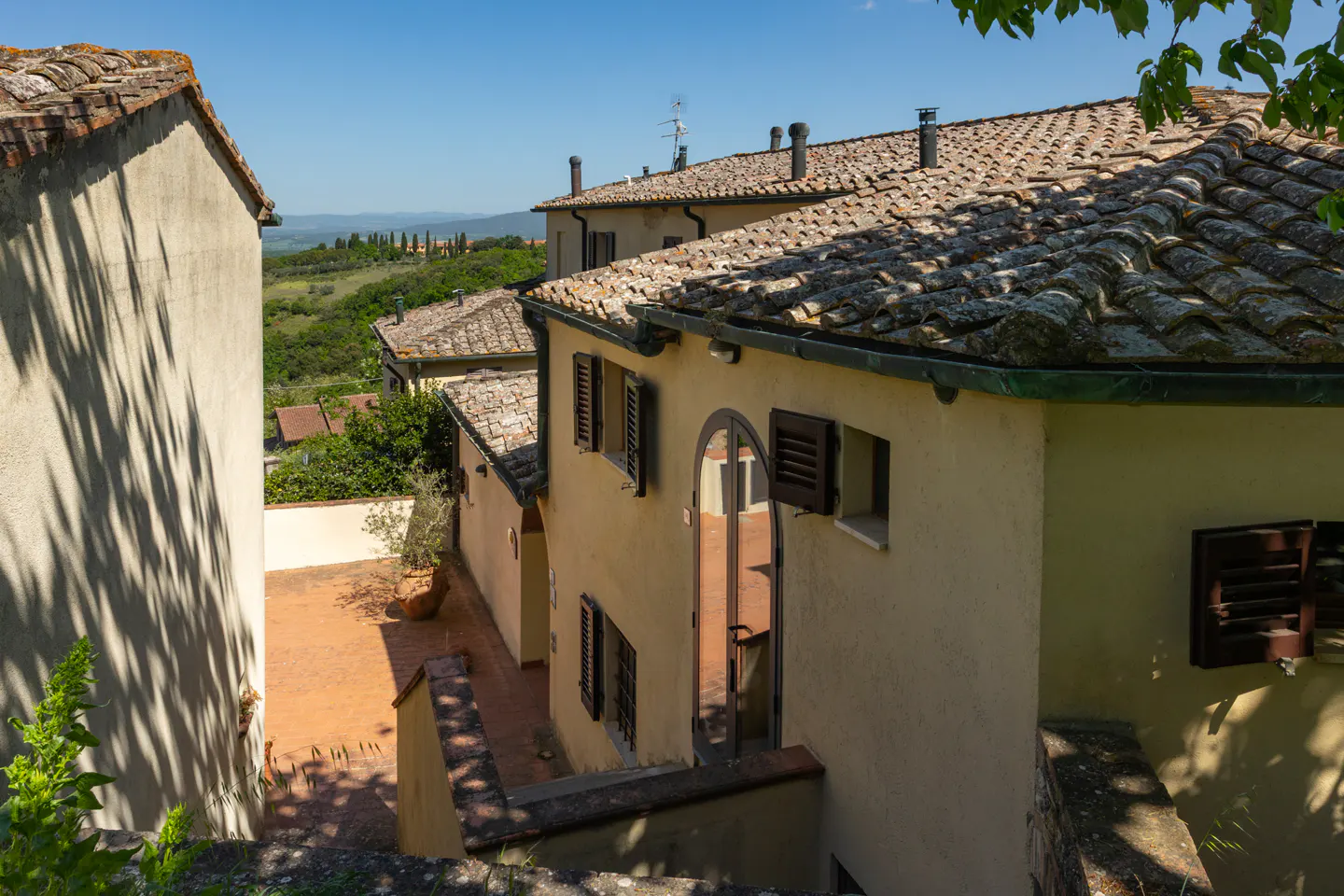 Tuscan-style building with terracotta roof tiles and brown shutters. A courtyard is visible, with green hills in the background.