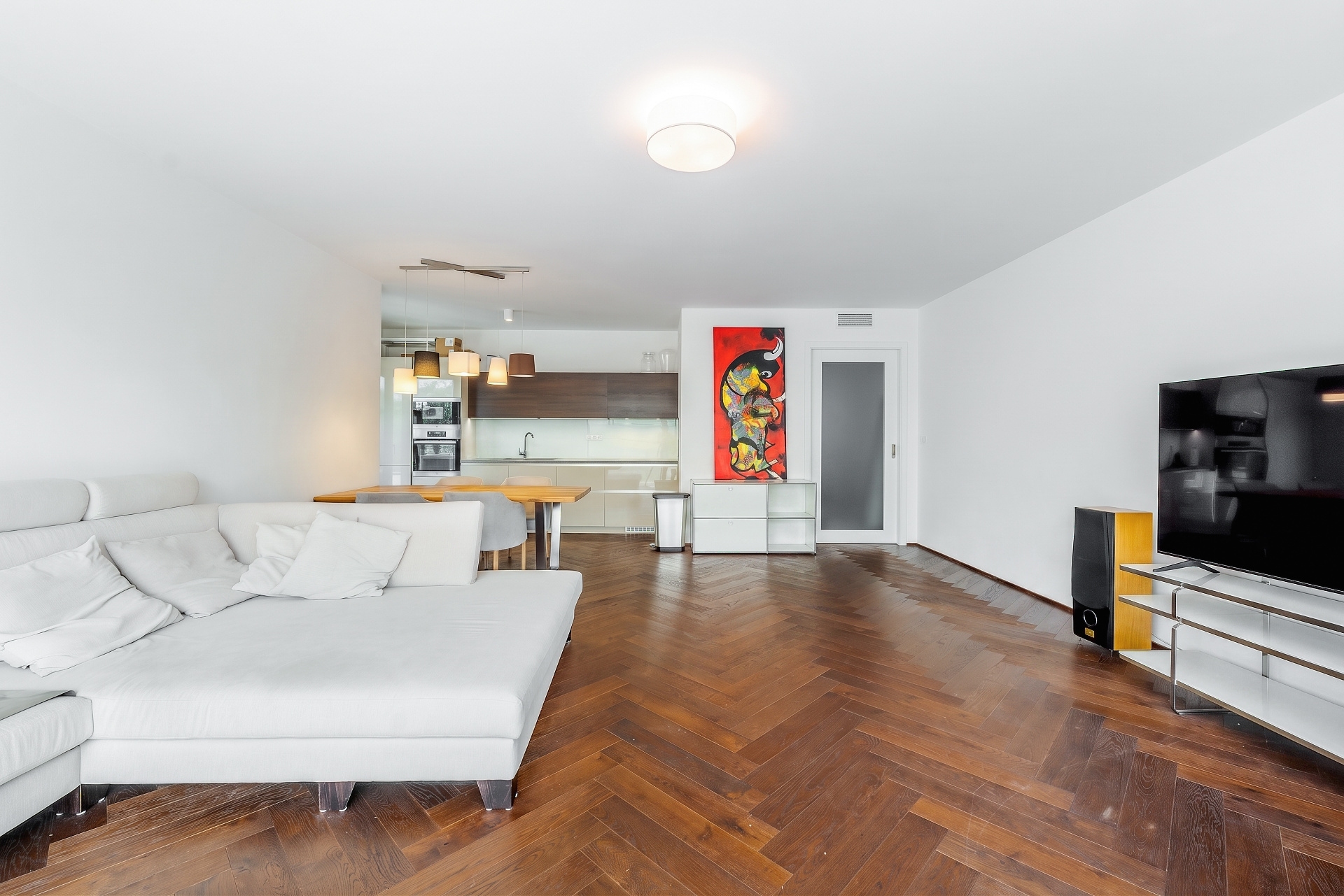 Modern living room with white sectional sofa, herringbone wood floors, TV, and open kitchen with dining area in background. [AI-generated text]