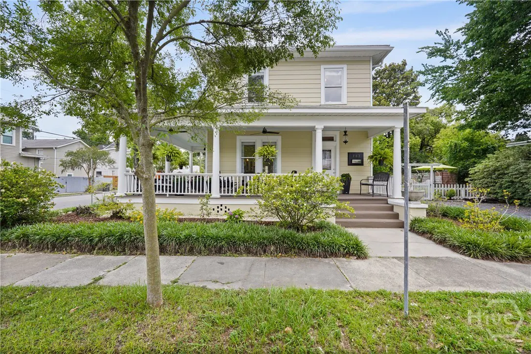 Two-story yellow house with a white porch and railing. Green lawn and trees surround the house.