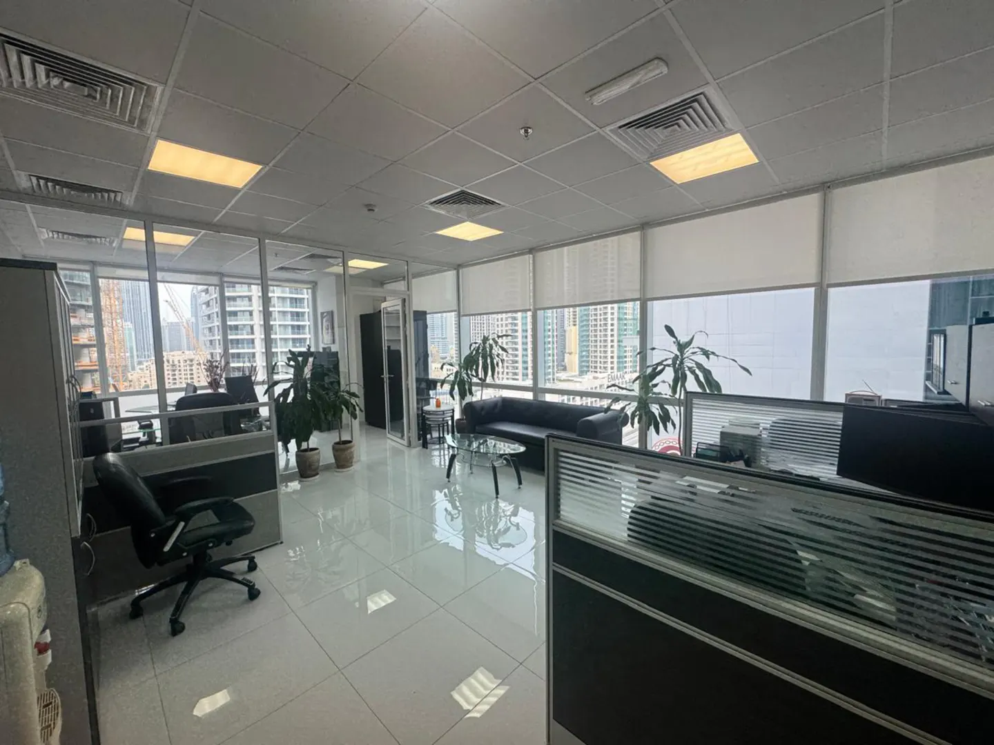 Bright office space with cubicles, a black sofa, and potted plants near large windows with blinds.