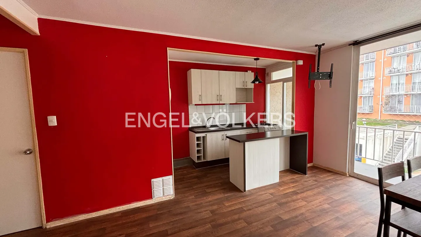 A modern apartment interior with red walls, wood floors, and a kitchen with light wood cabinets and a dark countertop island. A balcony door shows an exterior view.