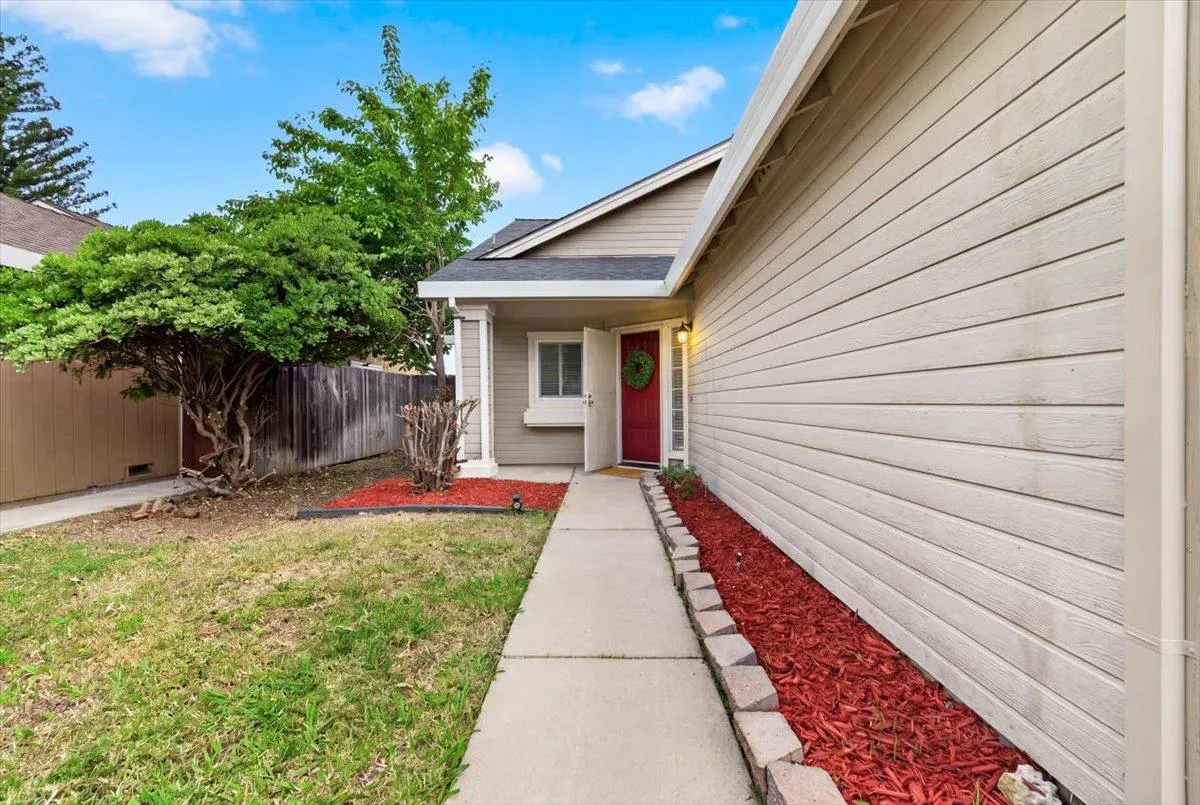 Tan house with a red door and green wreath. A concrete walkway leads to the entrance, with red mulch on either side. Blue sky above.