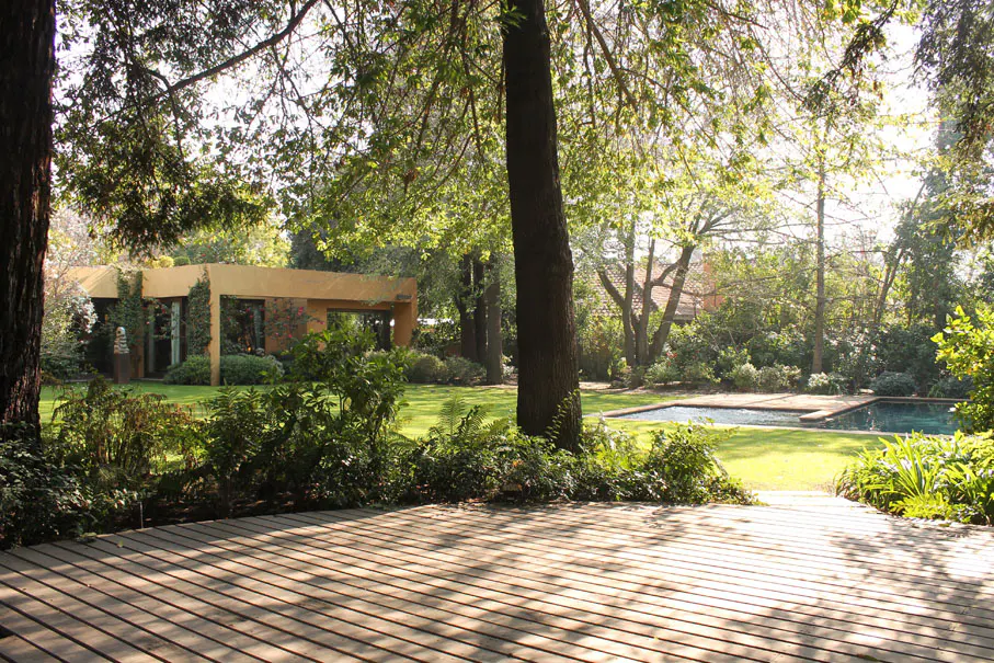 Wooden deck overlooking a green lawn, pool, and modern tan building surrounded by trees.