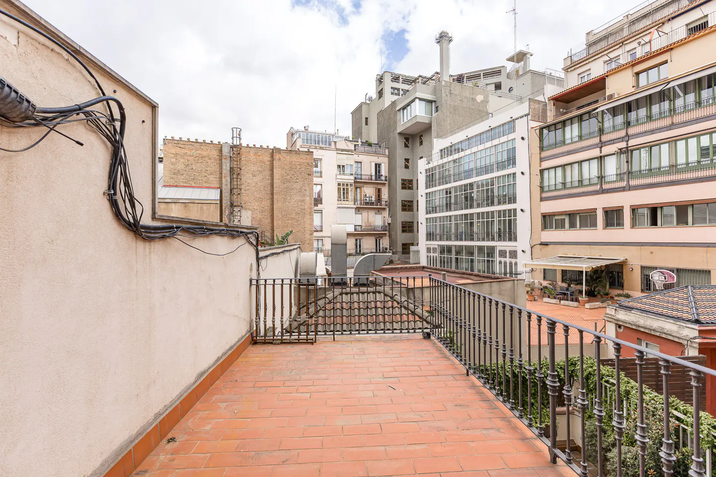 A rooftop terrace with red brick flooring and black iron railings, surrounded by buildings under a cloudy sky.