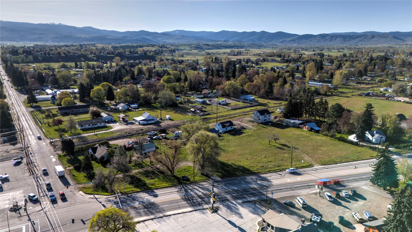 Aerial view of a small town with houses, trees, roads, and mountains in the background on a sunny day.