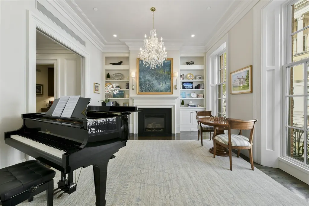 Bright living room with a black grand piano, fireplace, and a chess table by the window. A crystal chandelier hangs from the ceiling.