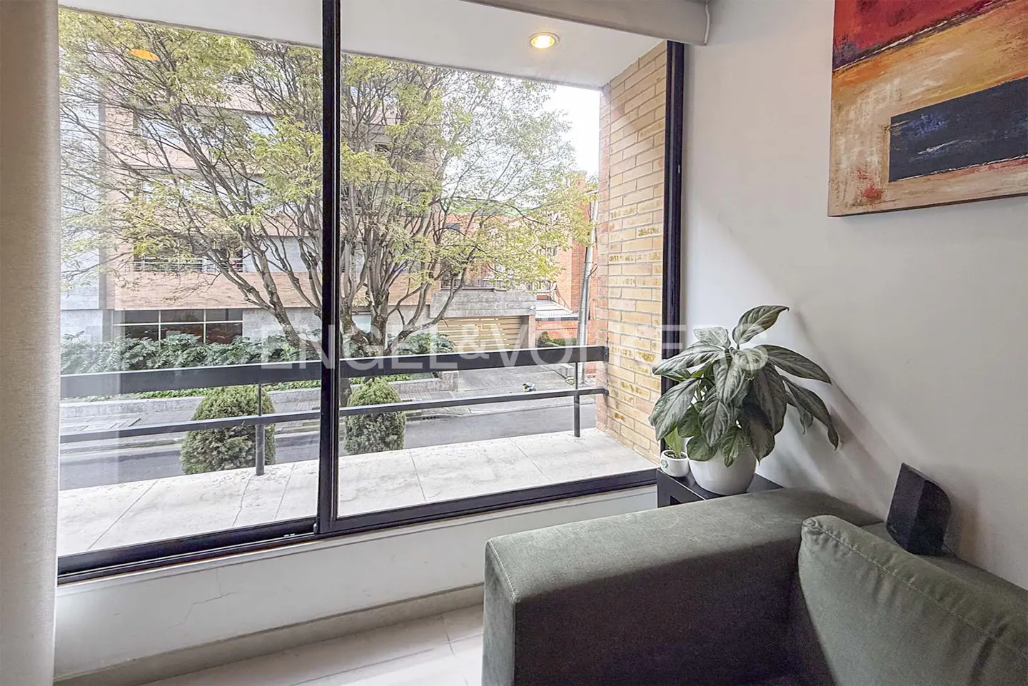 Living room view with a large window showing a city street with trees and buildings. A green sofa and plant are visible inside.