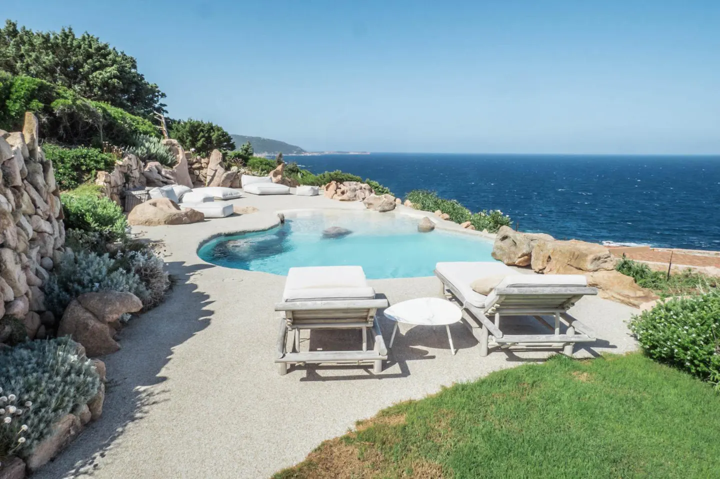 Infinity pool overlooking the ocean with lounge chairs on a stone patio, surrounded by rocks and lush coastal greenery.