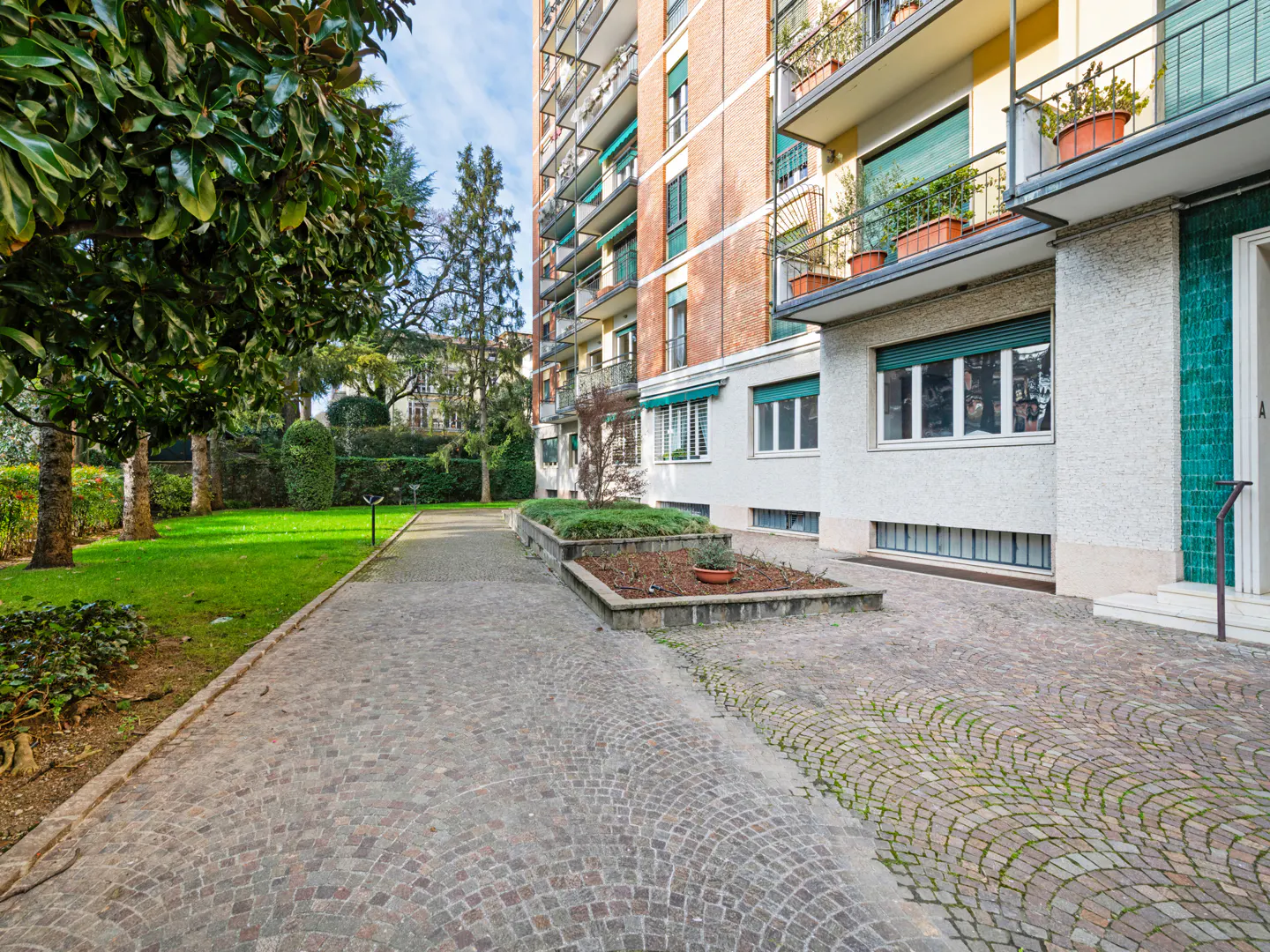 Exterior view of a brick apartment building with balconies, a cobblestone driveway, and a green lawn with trees.