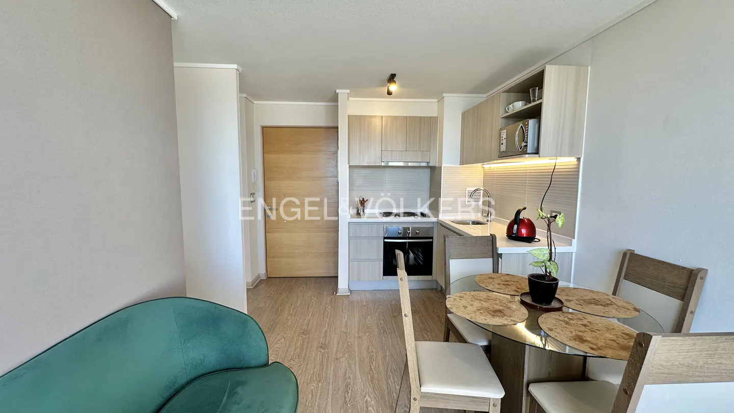 A compact apartment features a kitchen with light wood cabinets, a round glass table with four chairs, and a green velvet sofa.