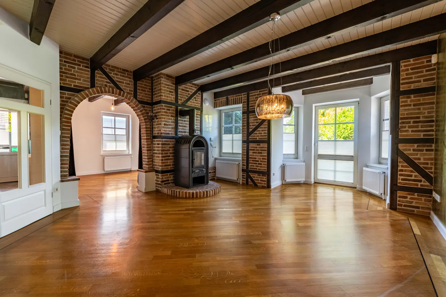 An empty living room with hardwood floors, exposed brick, and dark wood beams on the ceiling. A black wood-burning stove sits on a brick hearth.