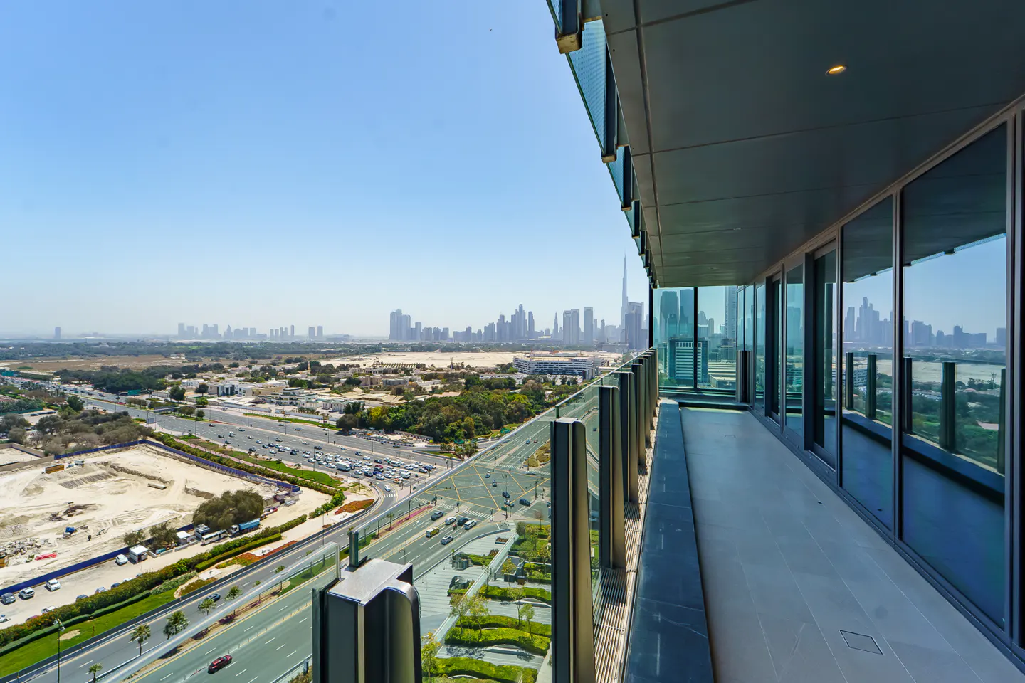 View from a high-rise balcony with glass railings overlooking Dubai's skyline on a sunny day. Cars drive on the road below.