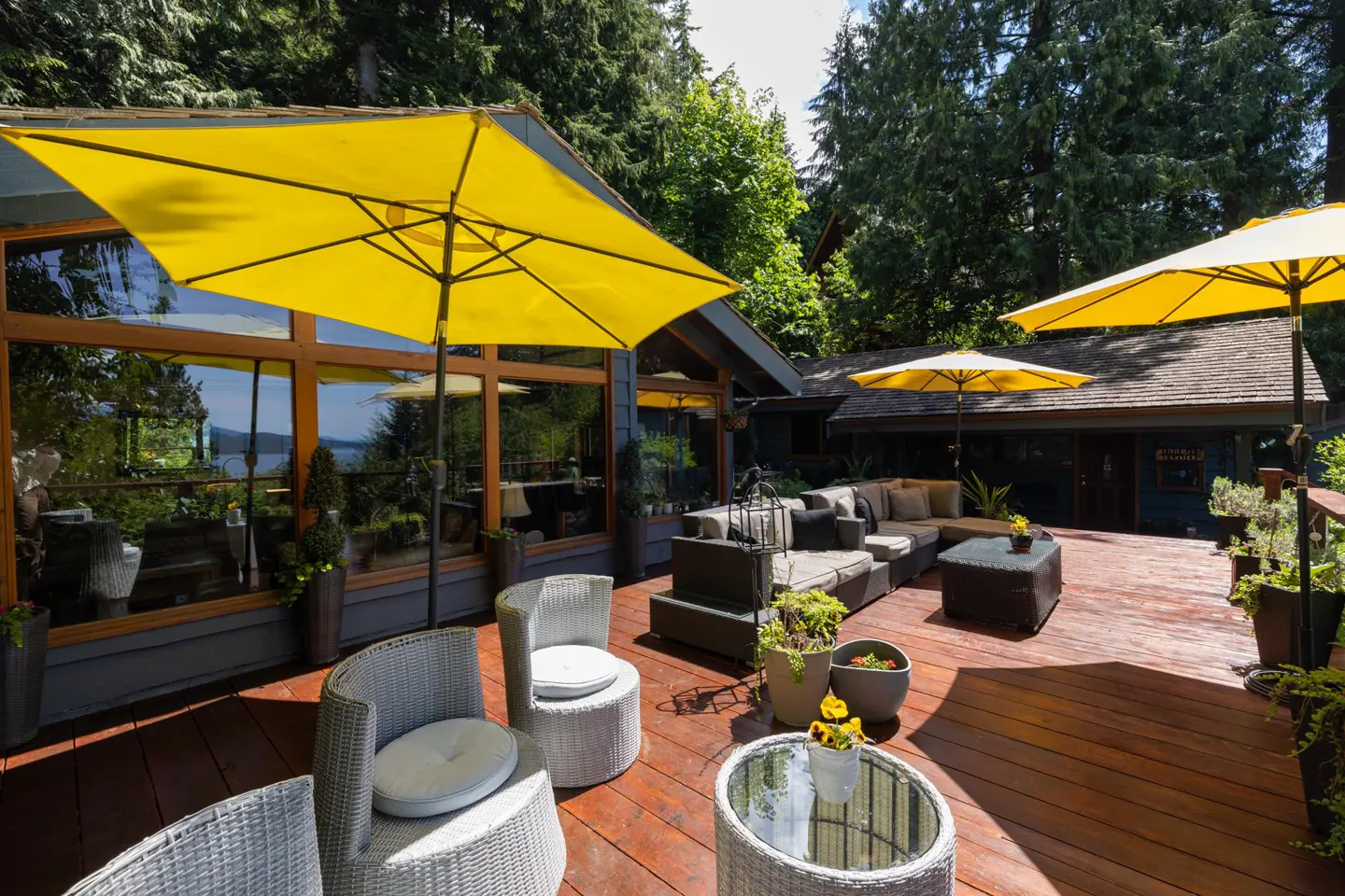 Outdoor deck with wicker furniture, yellow umbrellas, and potted plants. Large windows reflect a water view.