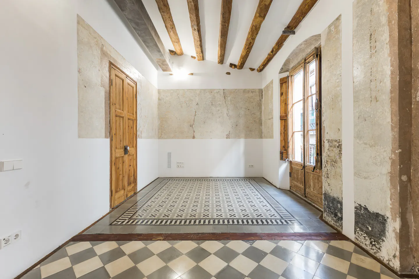 Empty room with wooden beams, door, and window. Walls are white and beige, with patterned tile floor.