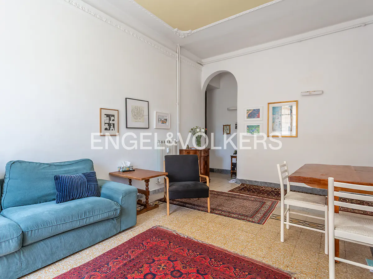 Bright living room with a blue sofa, black armchair, wooden dining table, and patterned rugs on a tiled floor.