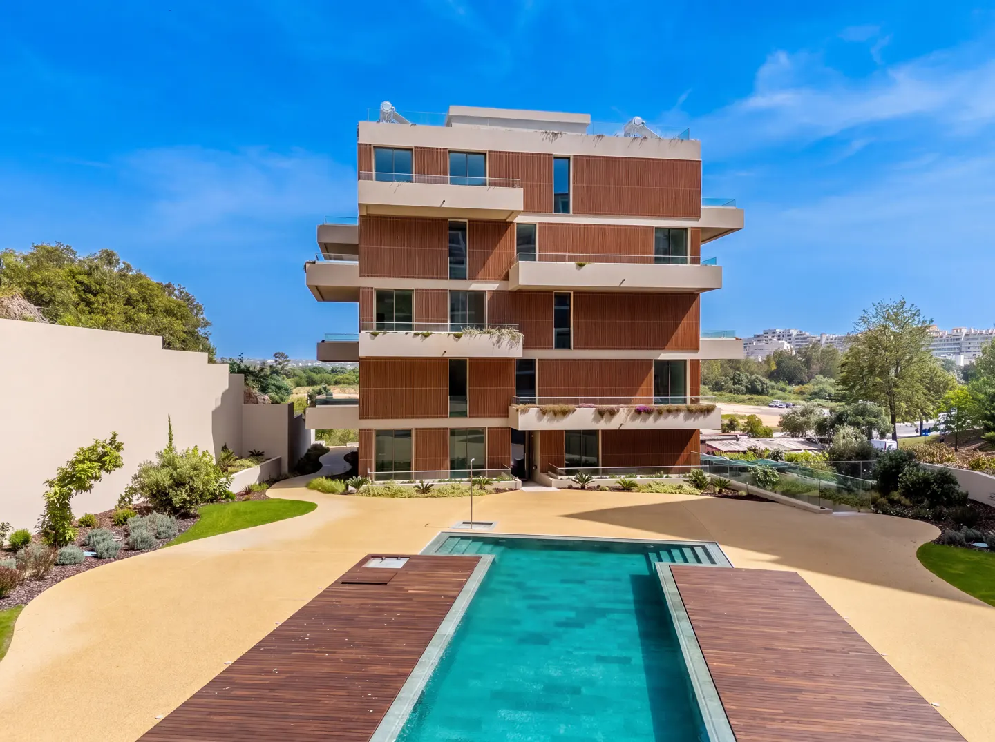 Modern apartment building with balconies overlooks a turquoise pool and tan courtyard under a bright blue sky.