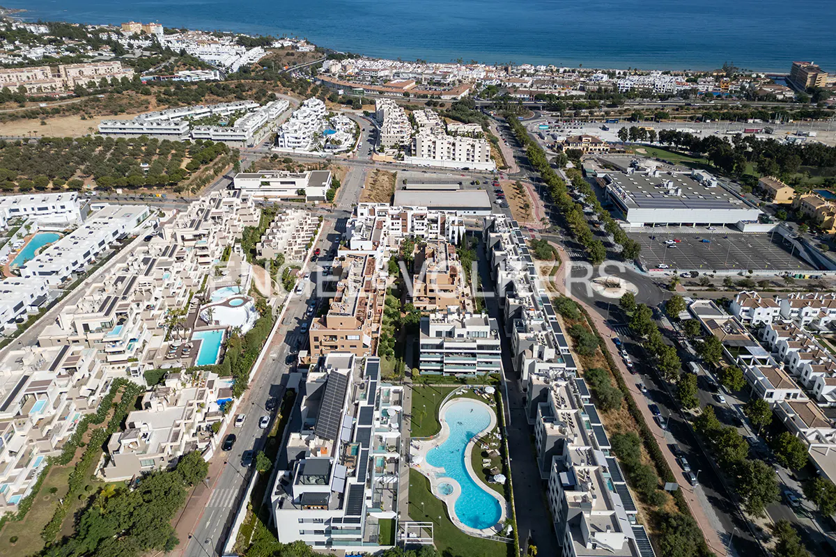 Aerial view of a coastal town with white buildings, pools, and the blue sea in the background.