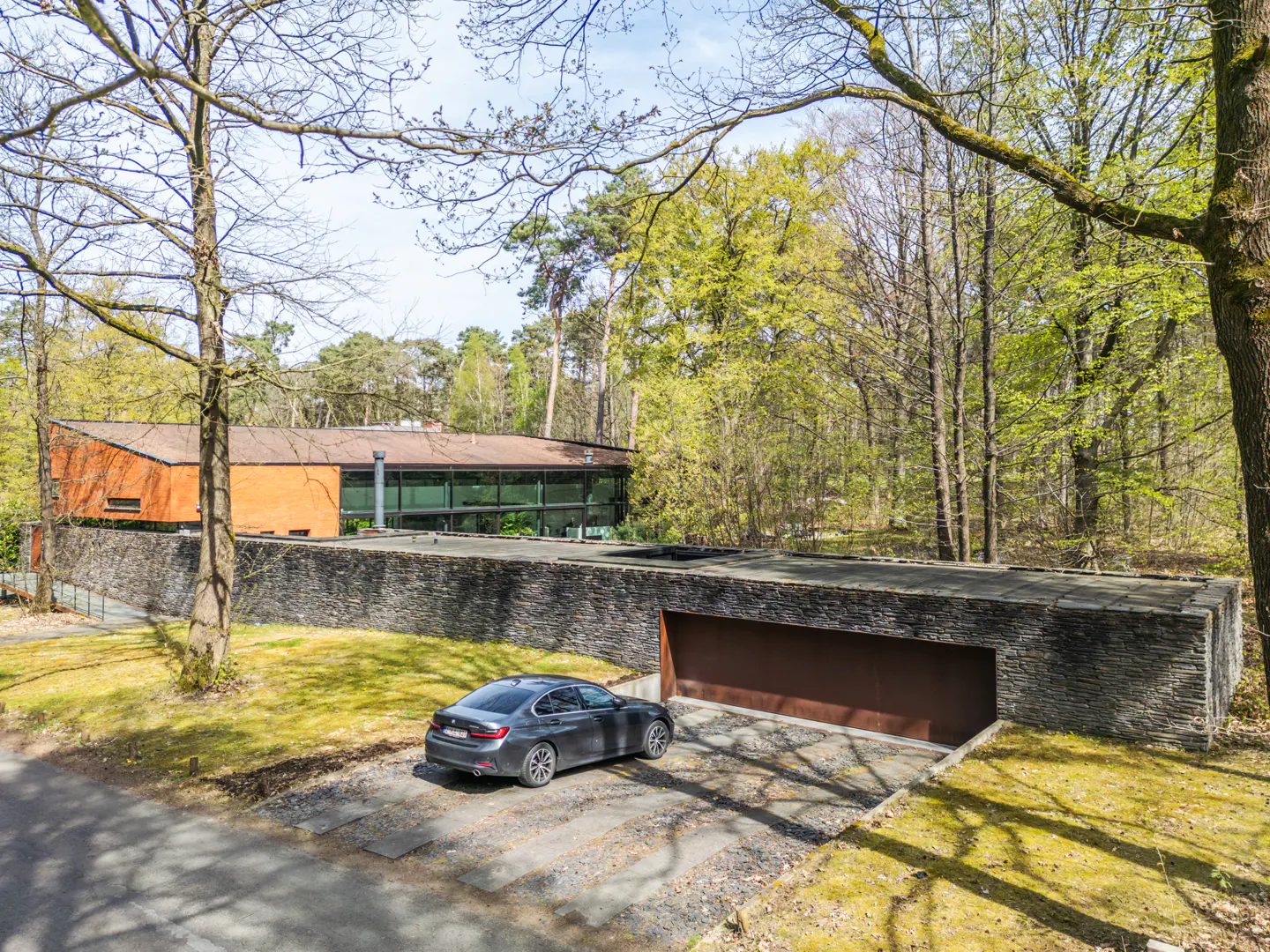 Exterior view of a modern brick and stone home with a gray car parked in front of a garage. Trees surround the property.