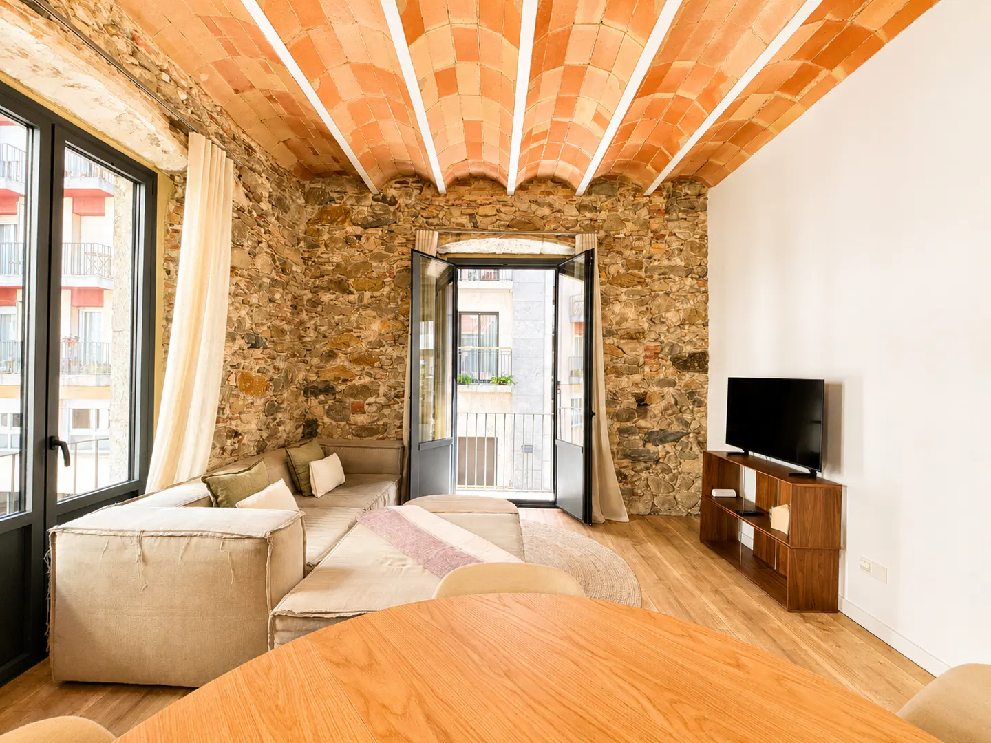 Bright living room with stone walls, brick ceiling, and wood floors. A large sectional sofa faces a TV. A round wood table is in the foreground.