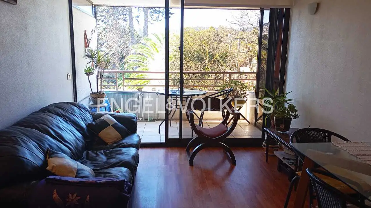 Living room with a black leather couch, wood floors, and sliding glass doors to a balcony with trees in the background.