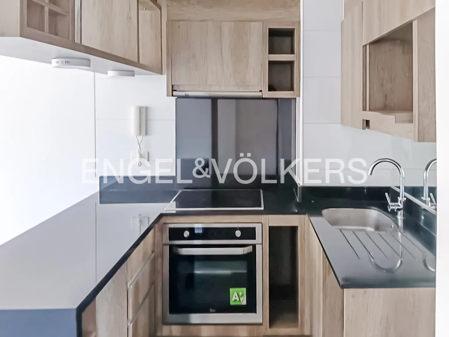 A modern kitchen with light wood cabinets, black countertops, and stainless steel appliances. The Engel & Völkers logo is visible.