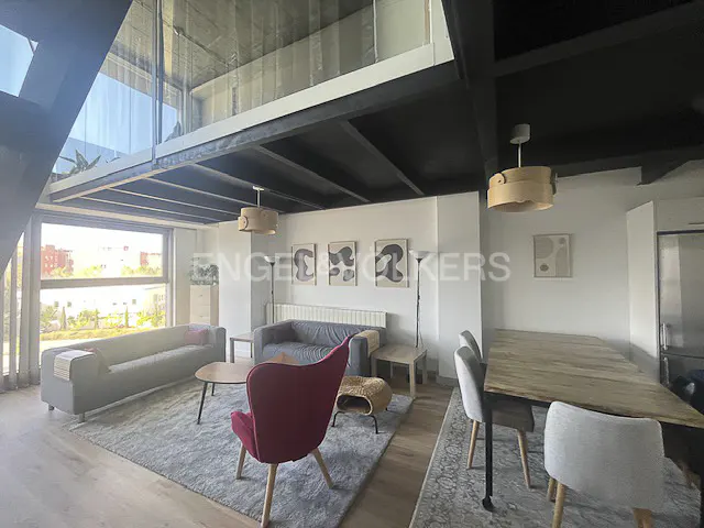 Modern open-plan living room with a red armchair, grey sofas, wooden dining table, and high ceilings with black beams.