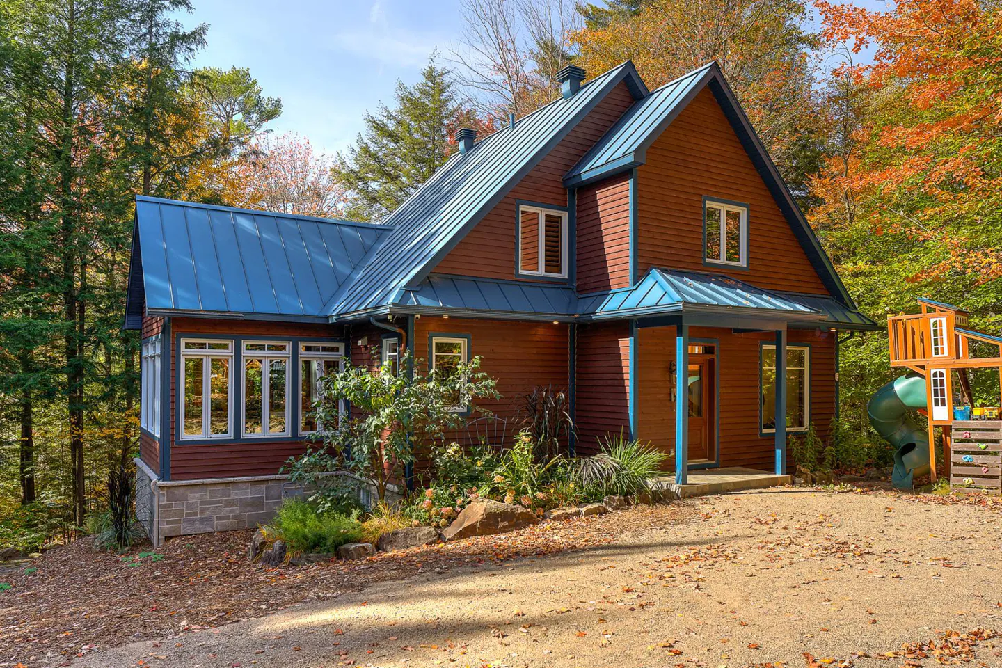 Exterior view of a two-story house with a blue roof and red siding, surrounded by trees in autumn. A playground is visible on the right.