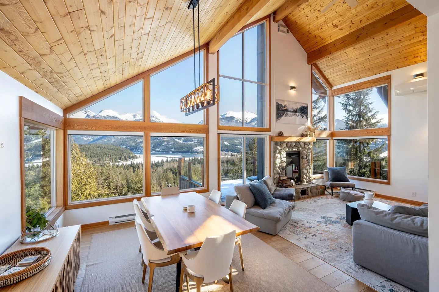 A bright living room with a wood ceiling, large windows with mountain views, a stone fireplace, and a dining table.