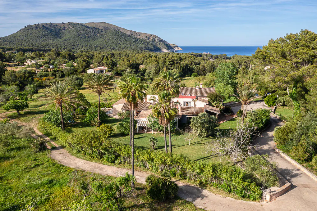 Aerial view of a large, tan house with a red roof, surrounded by palm trees and lush greenery, with a mountain and ocean in the background.
