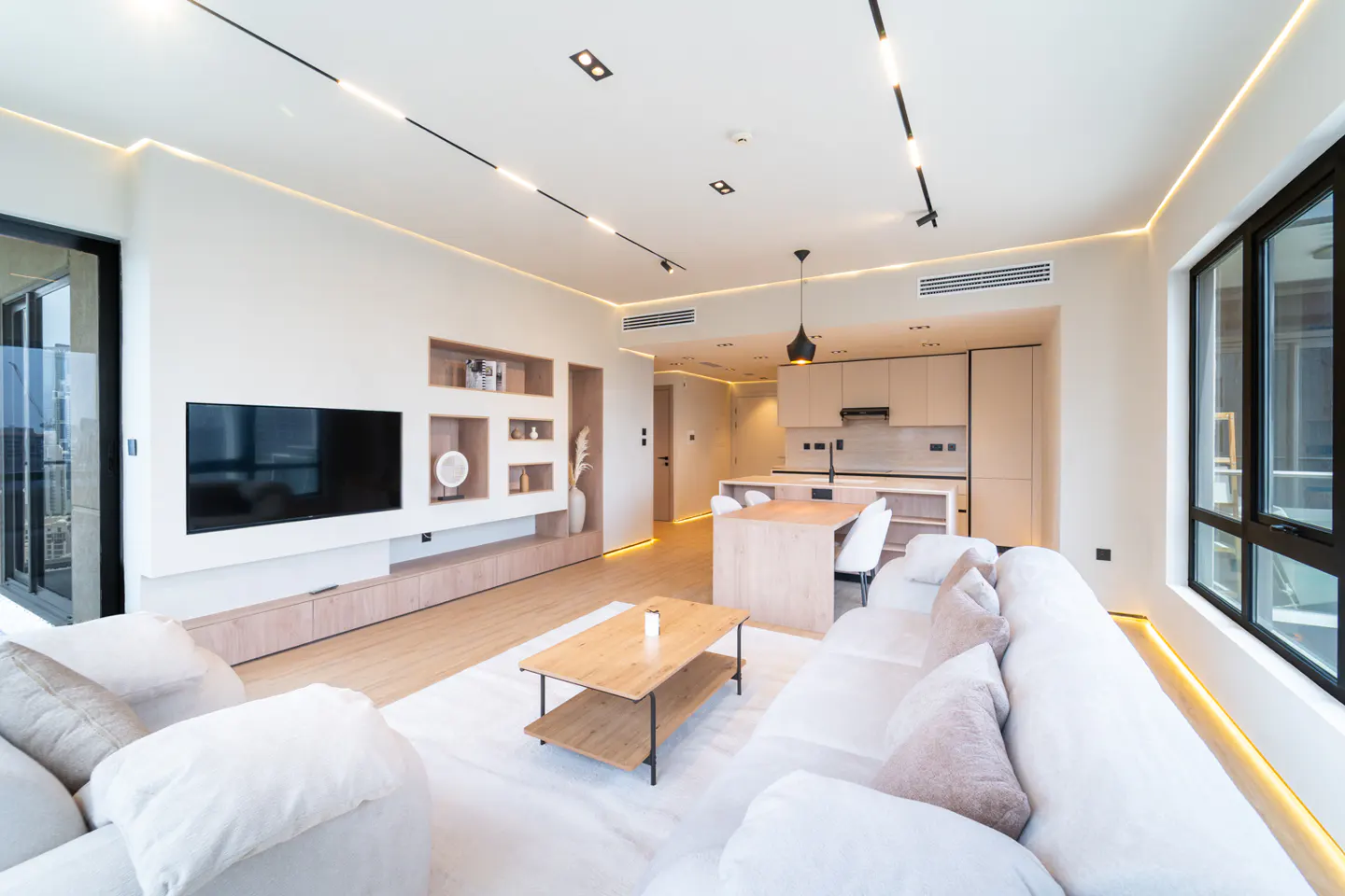 Bright, modern apartment interior with white sofa, wood coffee table, TV, and open kitchen. Large windows offer natural light.
