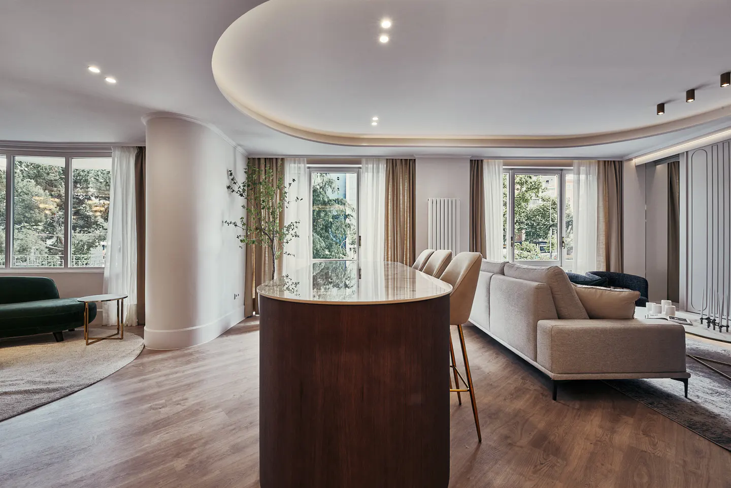 A modern living room with a curved ceiling, wood floors, and large windows. A dark wood island with bar stools separates the living area with a beige sofa and green couch.