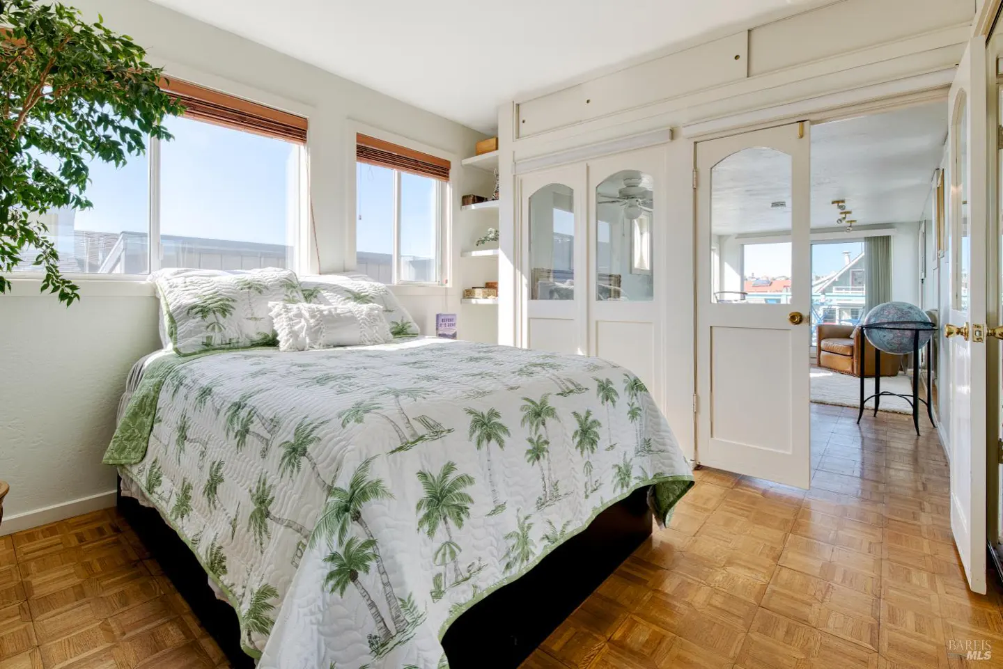 Bright bedroom with a palm tree patterned quilt, wood floors, and white walls. Windows let in natural light. Open doors lead to another room.