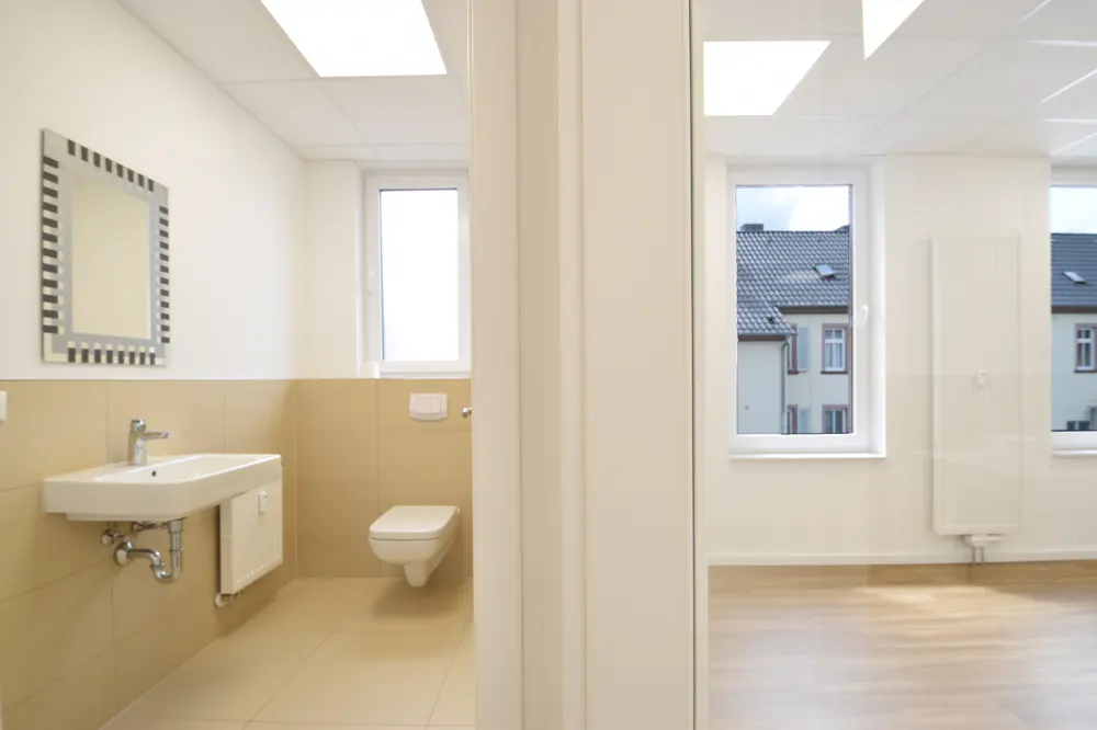 Modern bathroom with beige tiles, wall-mounted sink, toilet, and mirror, adjacent to a bright room with wood-look flooring.