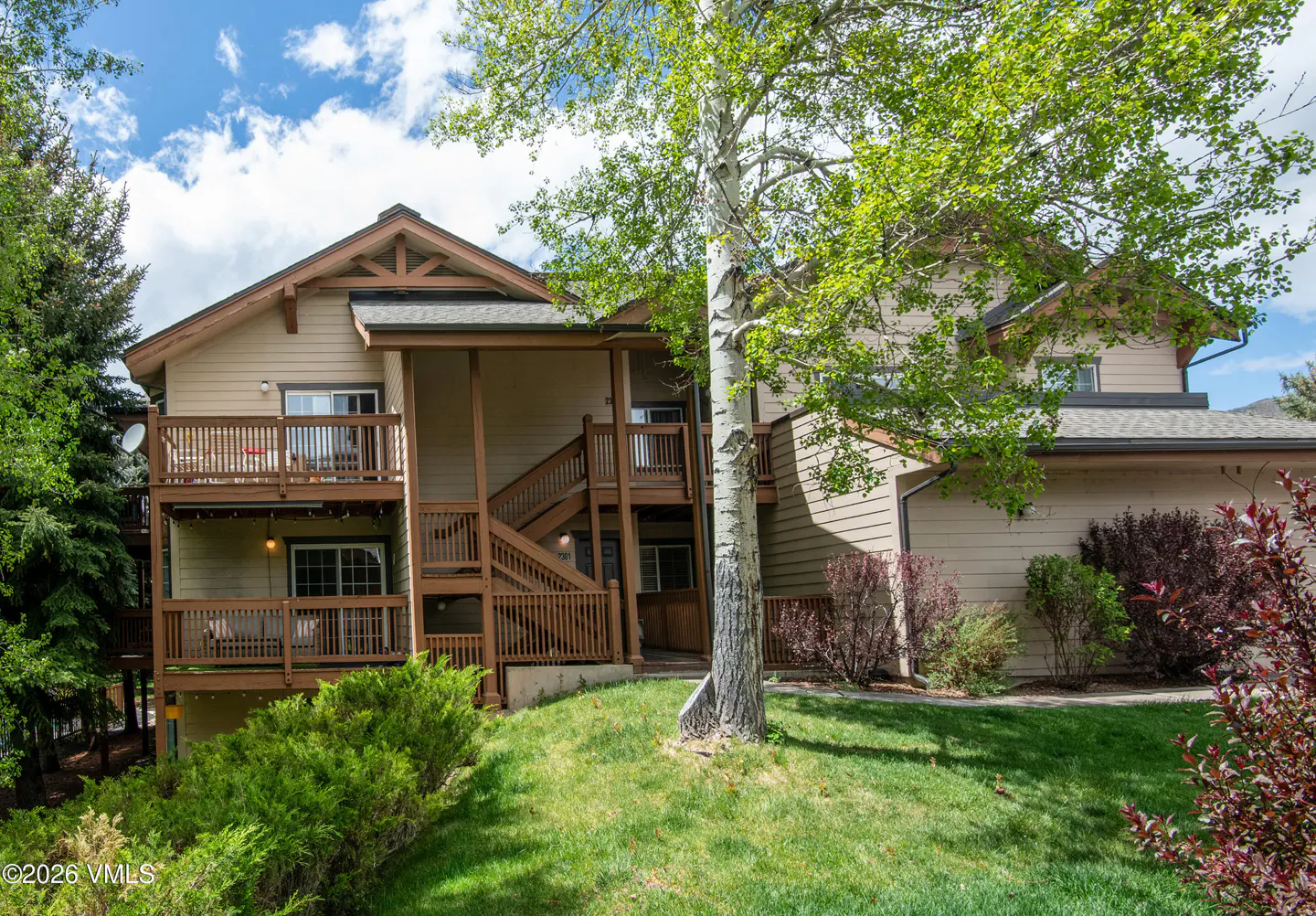 Two-story tan condo with brown trim and wooden balconies, surrounded by green grass and trees under a blue sky.