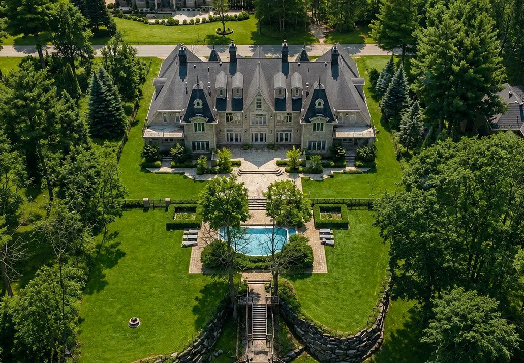 Aerial view of a large, gray stone mansion with a dark roof, surrounded by green trees and a manicured lawn with a blue pool.