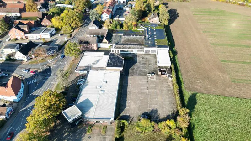 Aerial view of a commercial building with a large parking lot, surrounded by trees and a field.