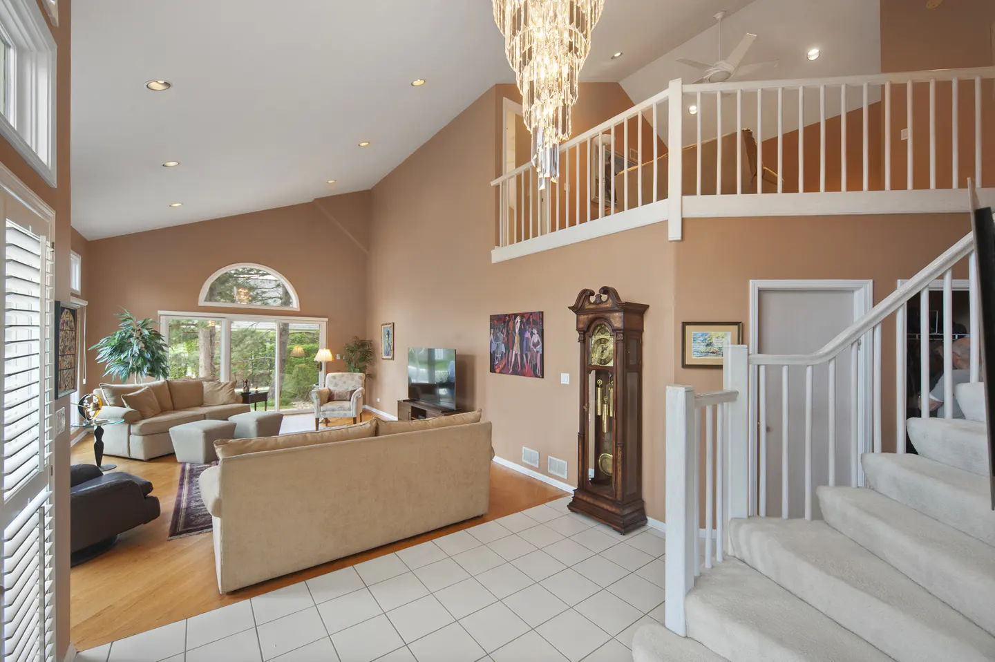 A spacious living room with tan walls, a chandelier, sofas, and a grandfather clock. A staircase leads to a second-floor balcony.