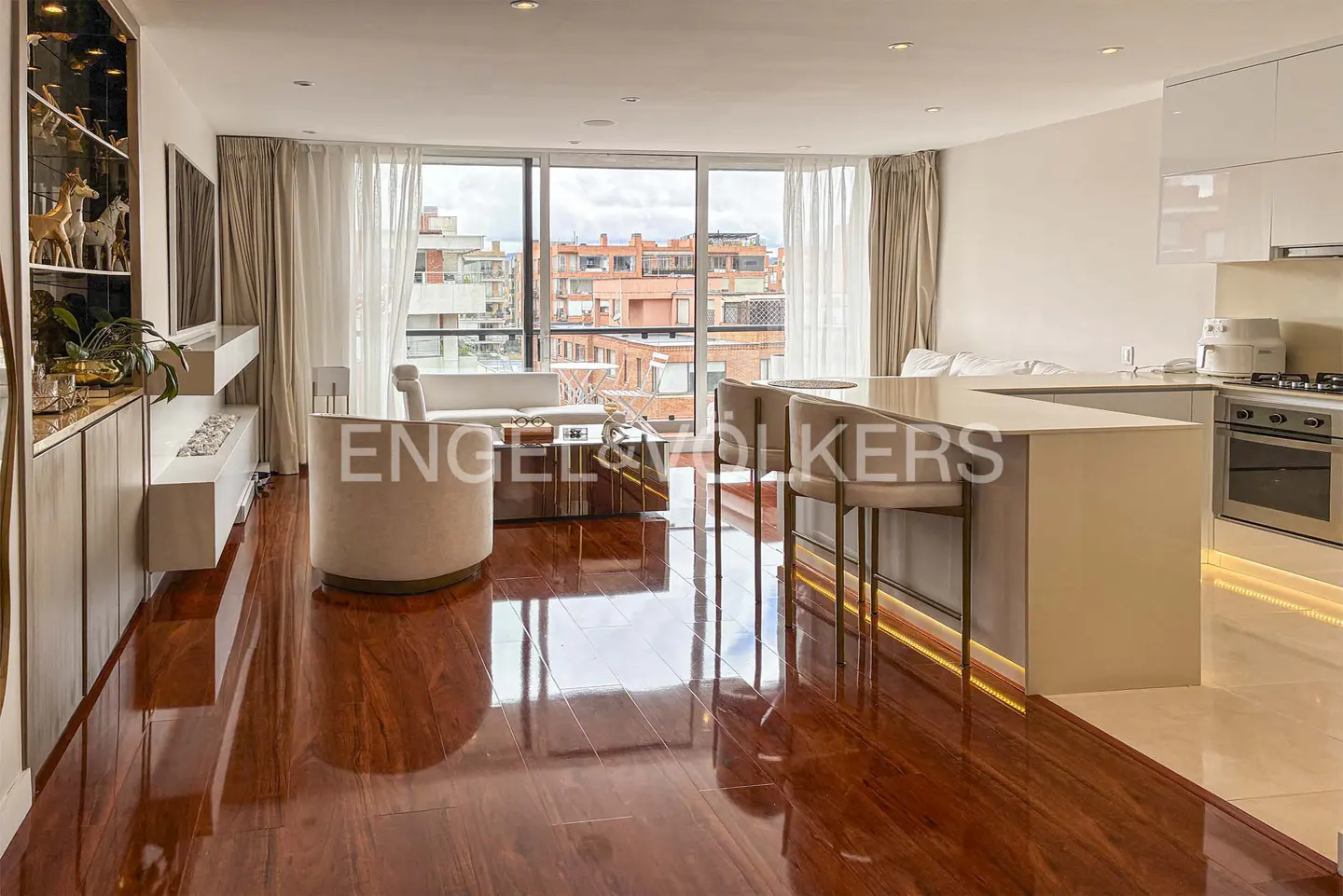 Open-concept living space with wood floors, white walls, and floor-to-ceiling windows. Kitchen island with bar stools and city view.