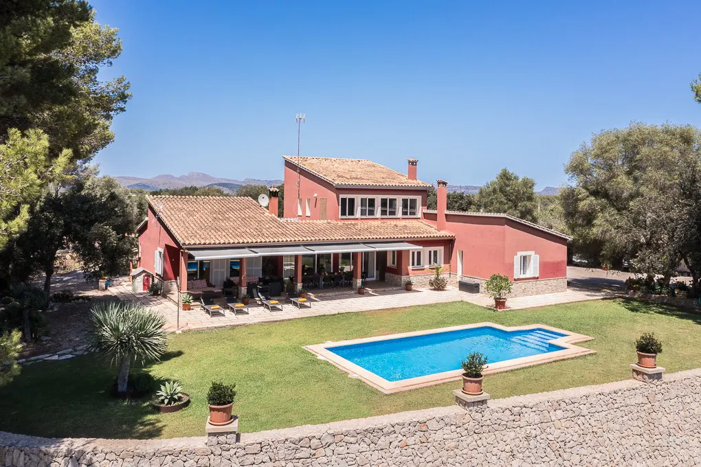 Aerial view of a red two-story house with a pool, green lawn, stone wall, and trees under a blue sky.
