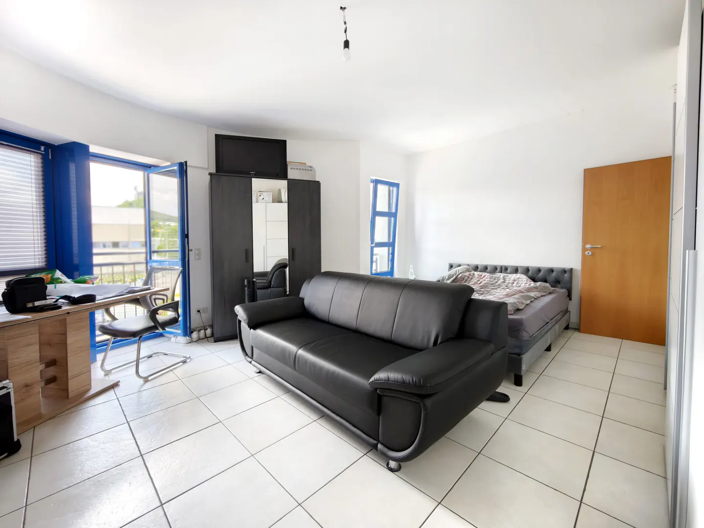Spacious studio apartment with white tile floors, a black leather sofa, a bed, a desk, and a balcony with blue frames.