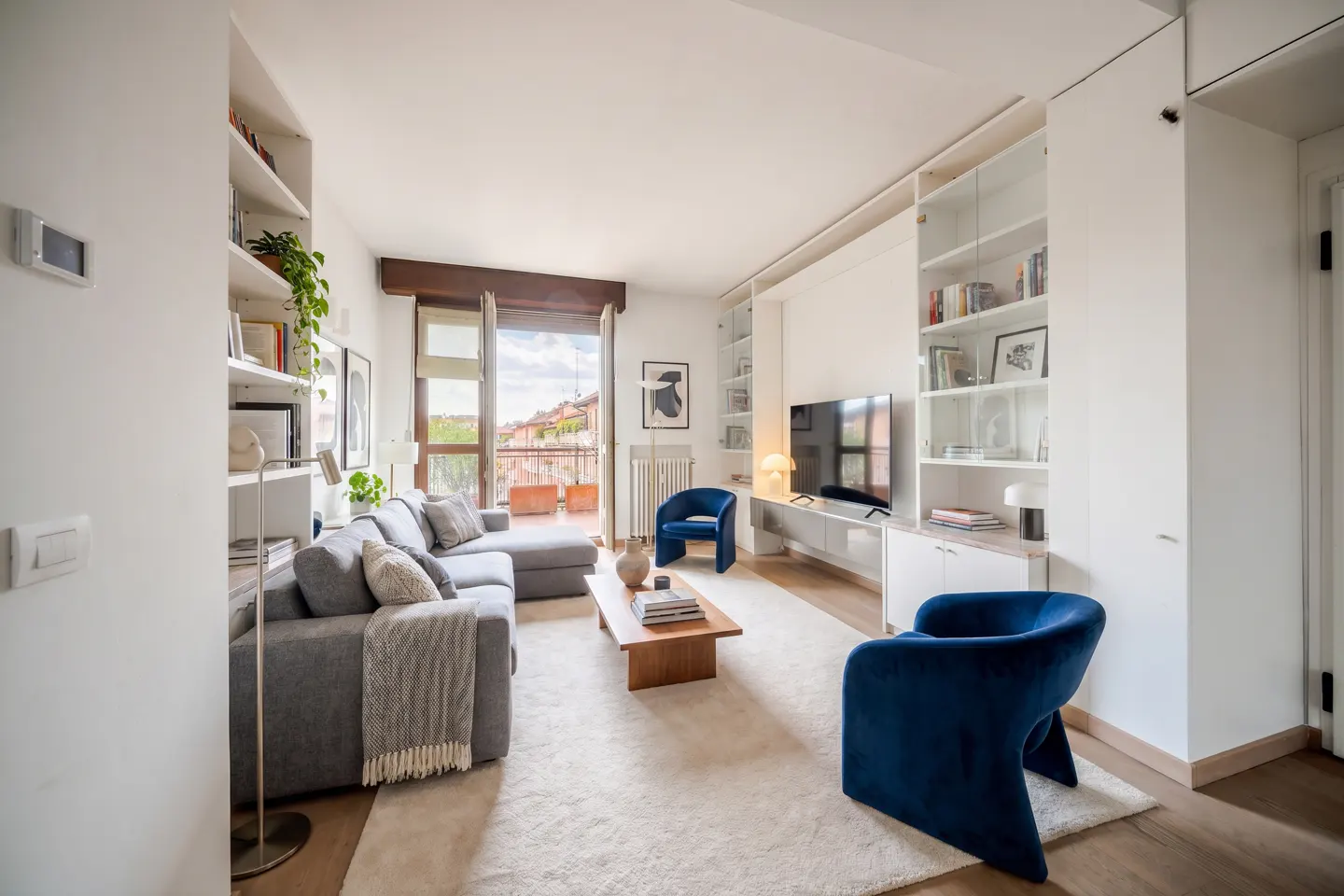 Bright living room with gray sofa, wood table, and blue chairs. Balcony doors open to a city view. White built-in shelves and TV unit.