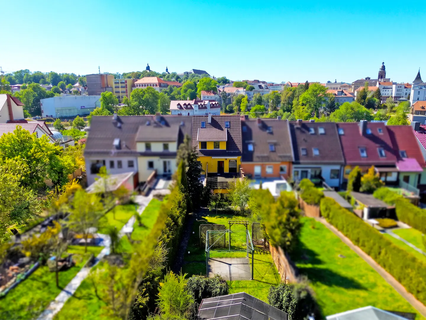 Aerial view of a row of townhouses with a yellow house in the center, green lawns, and a cityscape in the background.
