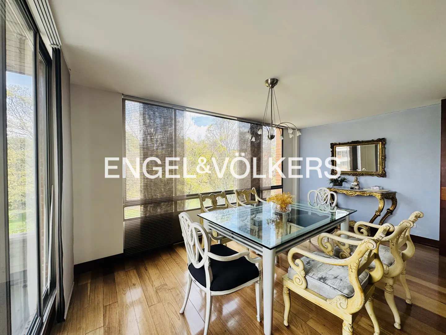 Dining room with glass table, white chairs, and wood floors. A gold mirror and console table are against a light blue wall.