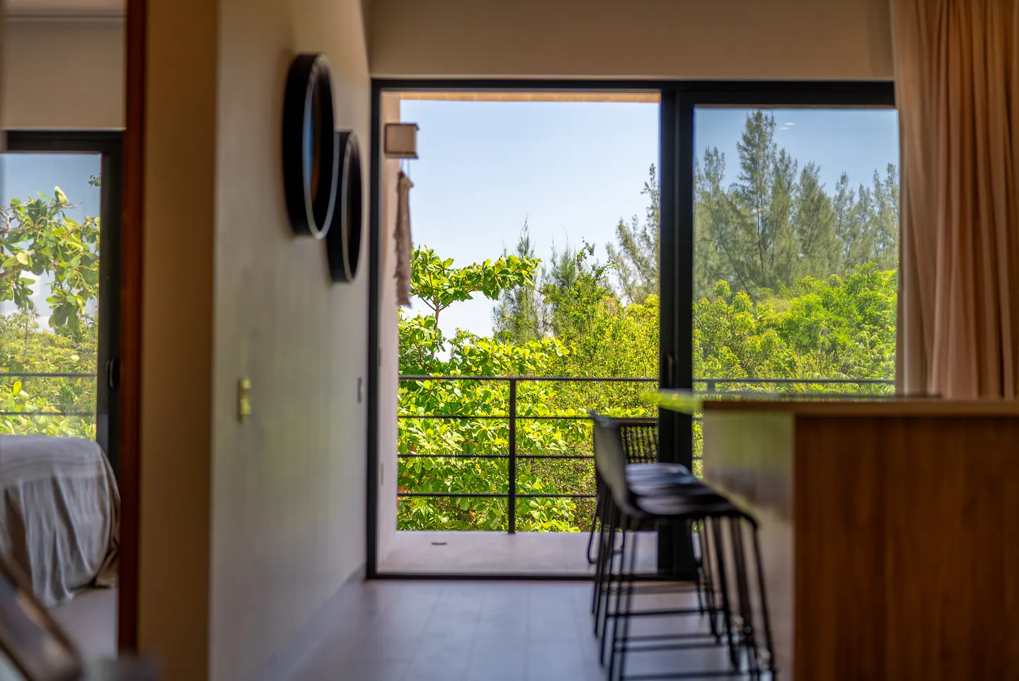 Interior view of a modern home with a balcony overlooking lush green trees. Black bar stools are visible near a wooden counter.
