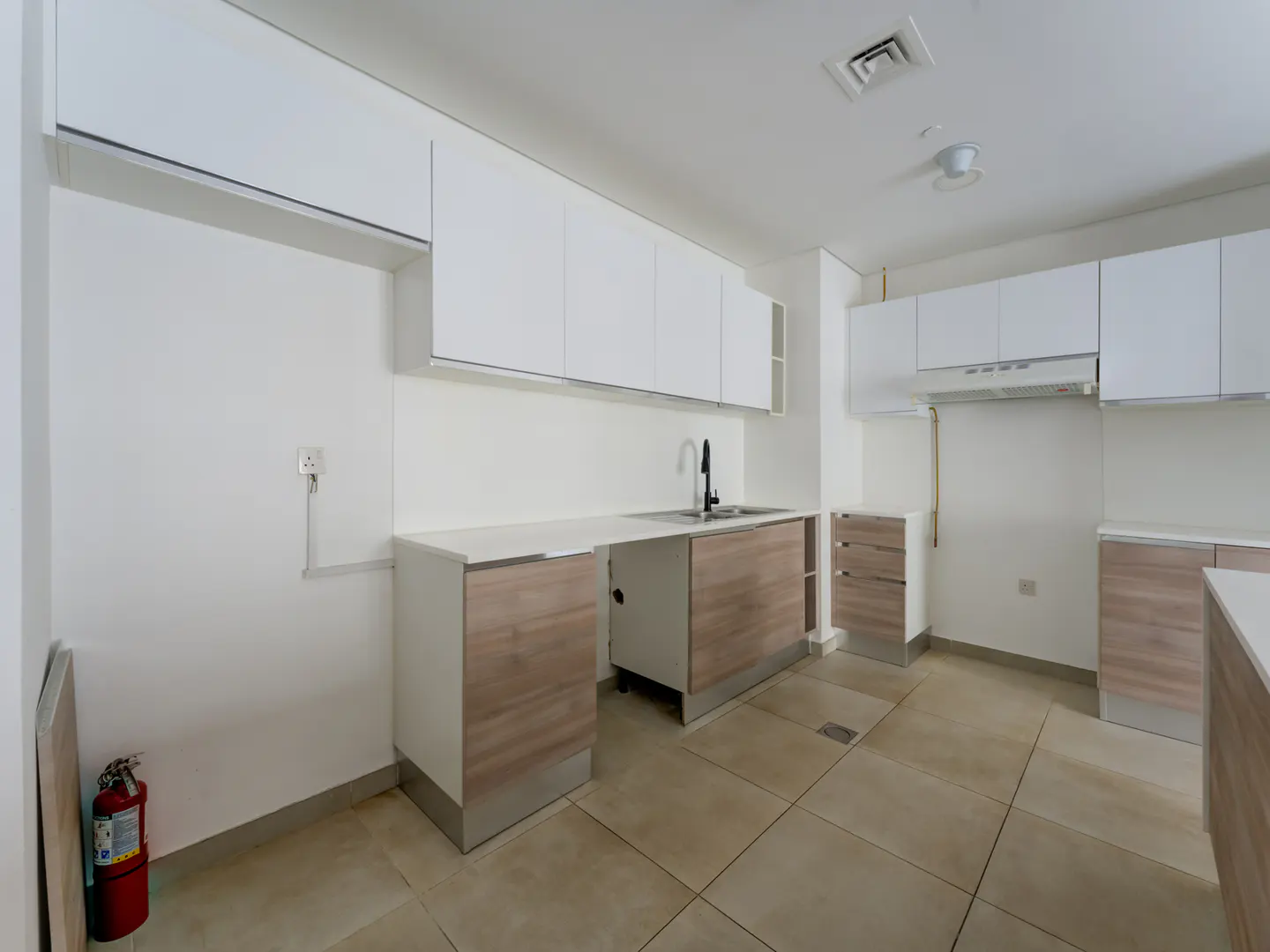 Bright kitchen with white cabinets, light wood lower cabinets, stainless steel sink, and beige tile floor. A red fire extinguisher is in the corner.