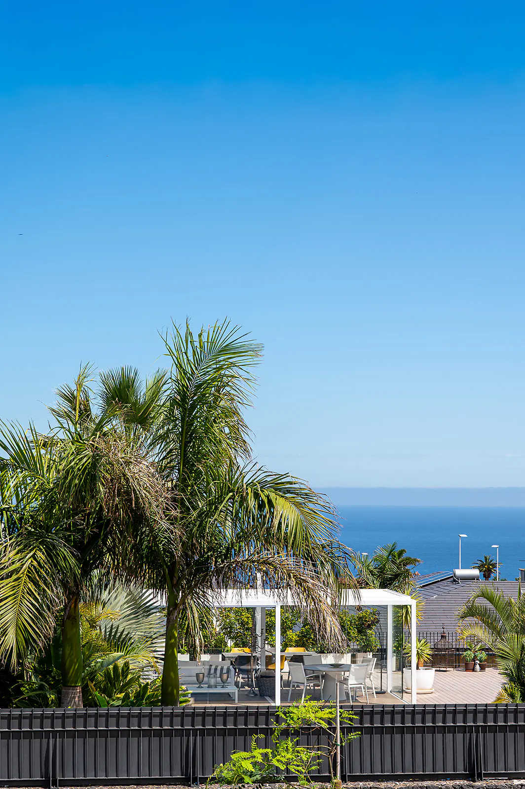 Outdoor patio with white pergola, table, and chairs, framed by palm trees and a black fence, with a blue sky and ocean view.