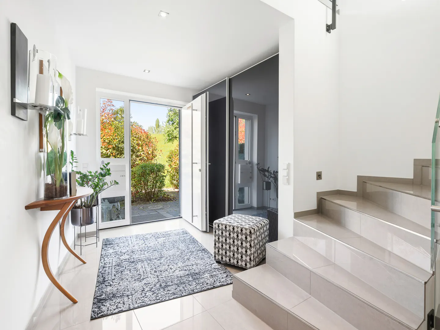 Bright foyer with open door to garden. A curved wood table holds plants. A patterned rug and ottoman sit near tiled stairs.