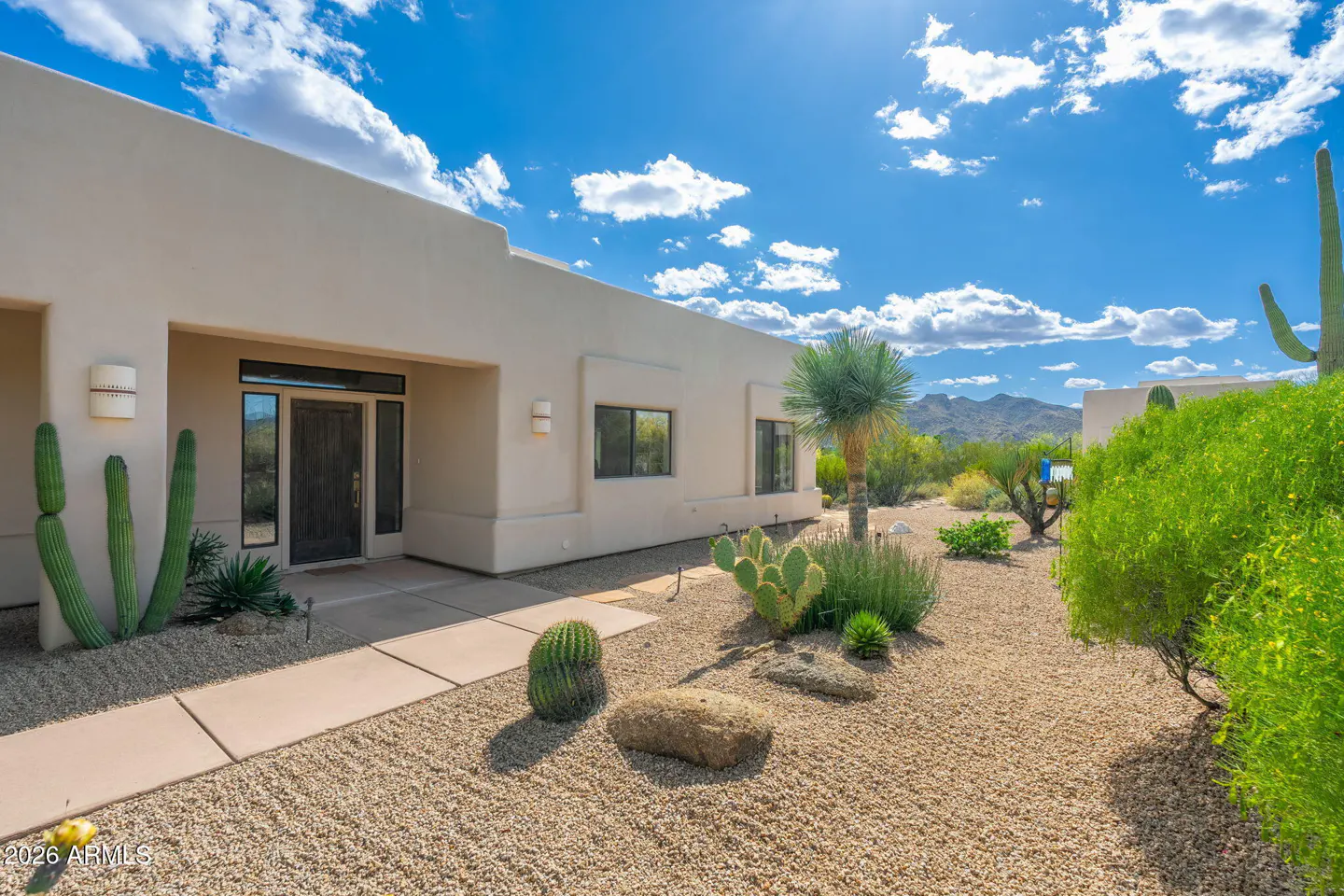 Beige southwestern home with a dark wood door, desert landscaping, and a blue sky with white clouds.