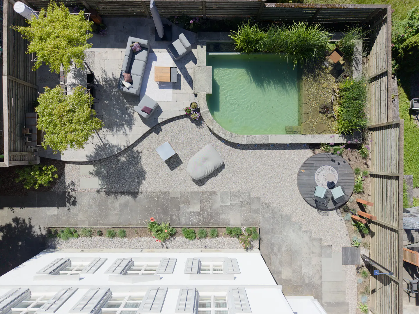 Aerial view of a backyard with a pool, patio furniture, and landscaping. The pool is surrounded by stone and plants. A gray sofa and chairs sit on a patio. A round table with chairs is near a wooden fence.