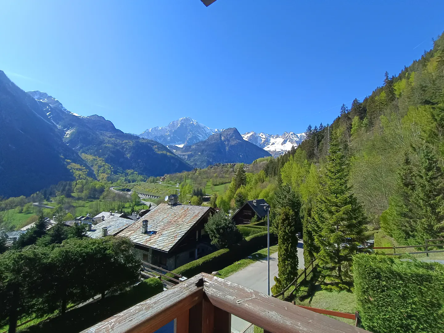 View from a balcony overlooking a valley with houses, green trees, and snow-capped mountains under a clear blue sky.